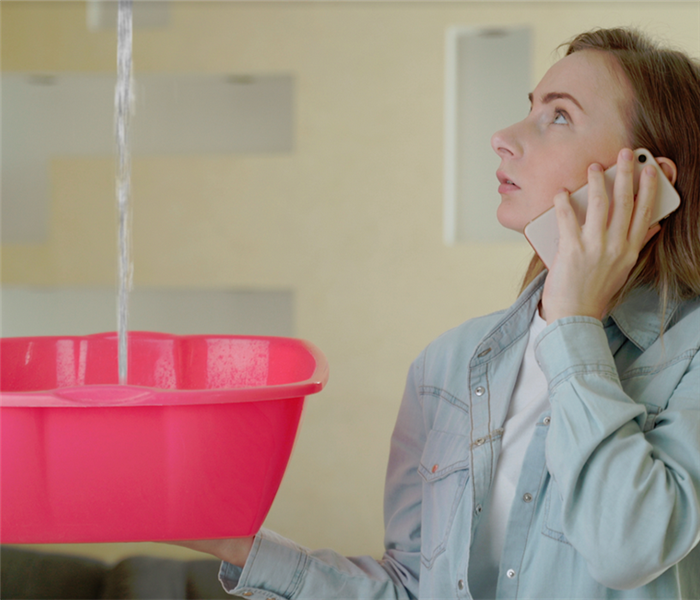 a woman on the phone holding a bucket to catch water falling from her ceiling